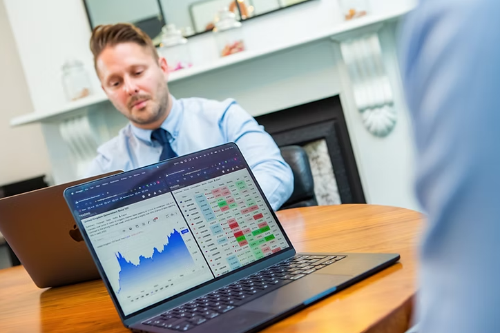 A man in a blue shirt and tie is sitting at a wooden table with a laptop, engaged in a meeting. In the foreground, another open laptop displays financial data and stock charts. 