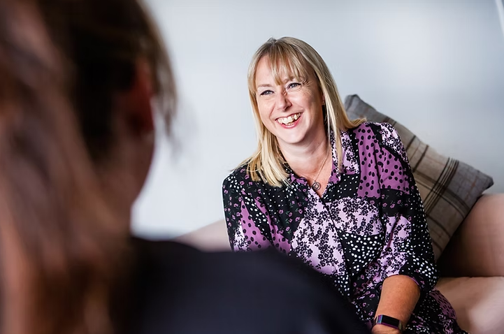 A woman with blonde hair is sitting on a sofa, smiling warmly during a conversation. She is wearing a patterned purple and black blouse, and the setting appears relaxed and informal, with a cushion behind her and another person slightly out of focus in the foreground.