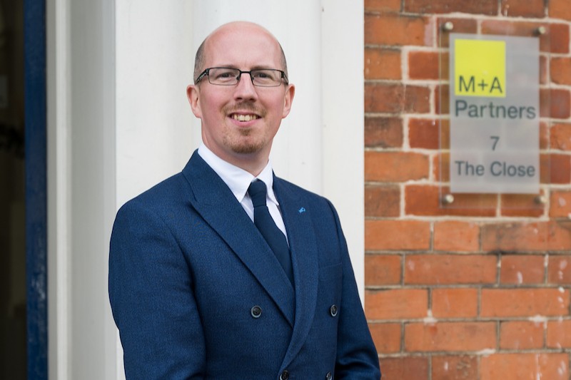 Sam Holloway dressed in a navy suit standing outside the M+A Partners Norwich office