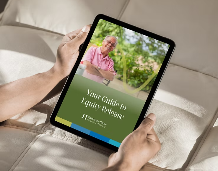 A person is holding a tablet while sitting on a cream-colored sofa. The screen displays a digital brochure titled "Your Guide to Equity Release" by Brancaster House Financial Planning. The cover features an elderly man in a pink polo shirt smiling and sitting outdoors in a lush green garden setting.