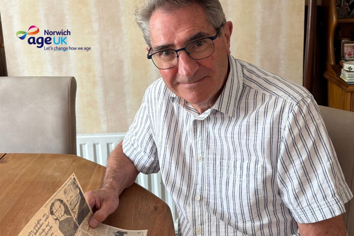 An older man wearing glasses and a striped short-sleeve shirt sits at a wooden table, holding an old newspaper clipping. He looks toward the camera with a gentle smile. The Age UK Norwich logo is visible in the top left corner of the image.