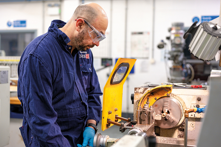 A gentleman in a blue jumpsuit wearing safety glasses works on a metal machine while wearing blue plastic gloves. He is looking down at the machine. 