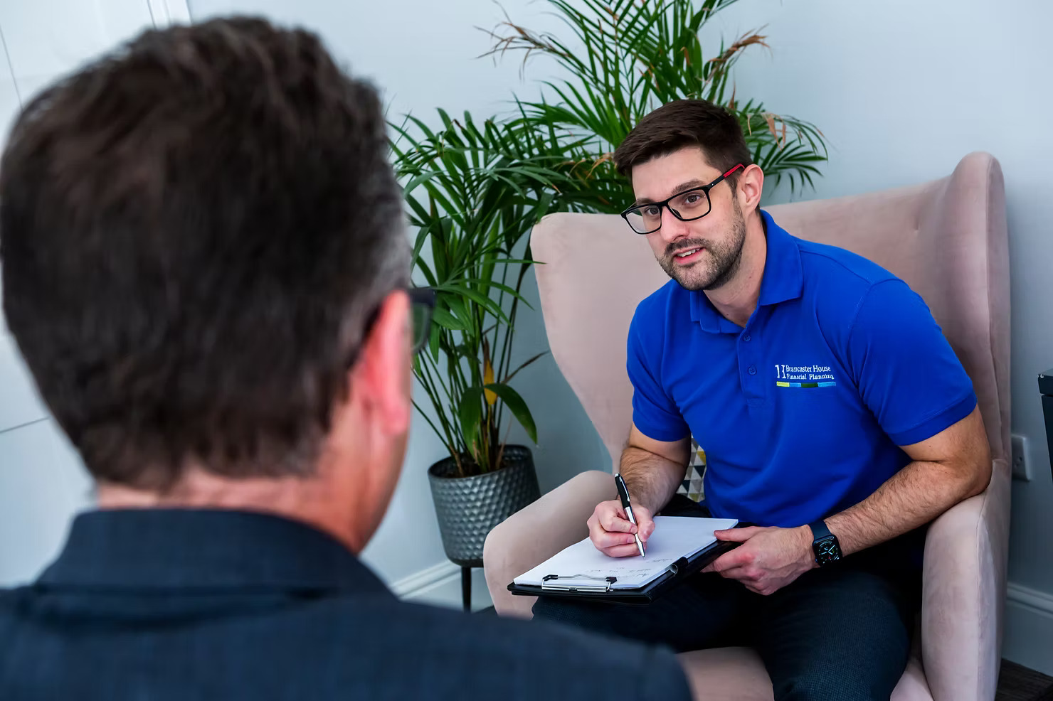 Man in blue shirt takes notes during a professional meeting with another person, seated in an office with a potted plant in the background.