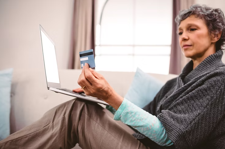 a short haired lady resting a laptop on her knees looking at a credit card