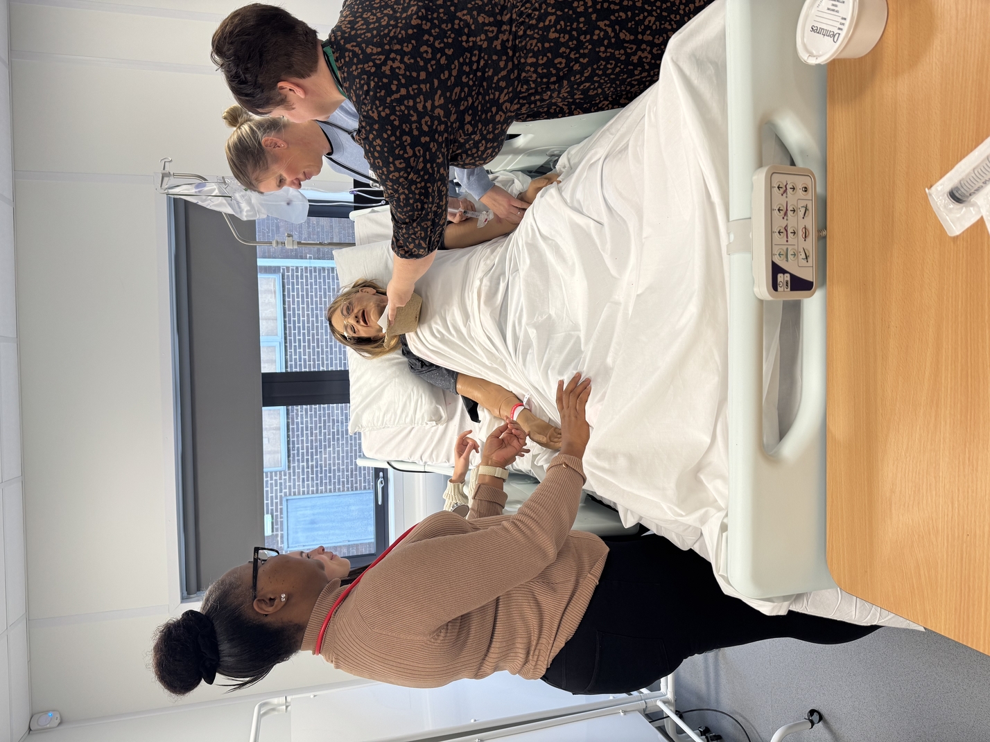 There are four people with their backs turned to the camera, roleplaying with a simulated mannequin who is being a patient 