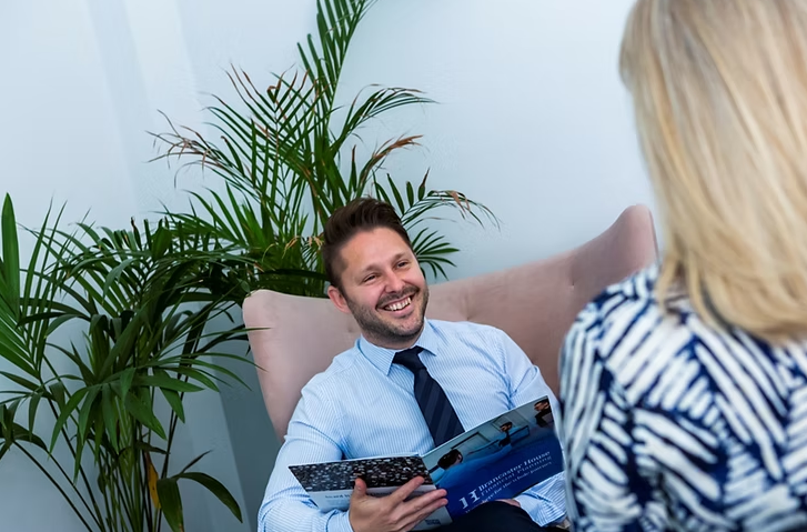 A man in a shirt and tie sits smiling in a chair, holding a brochure, while talking to a woman. Plants are in the background.
