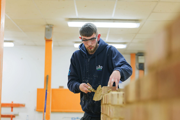 There is a man looking down and placing bricks onto a wall, he is wearing a blue hoody and wearing see through safety 
