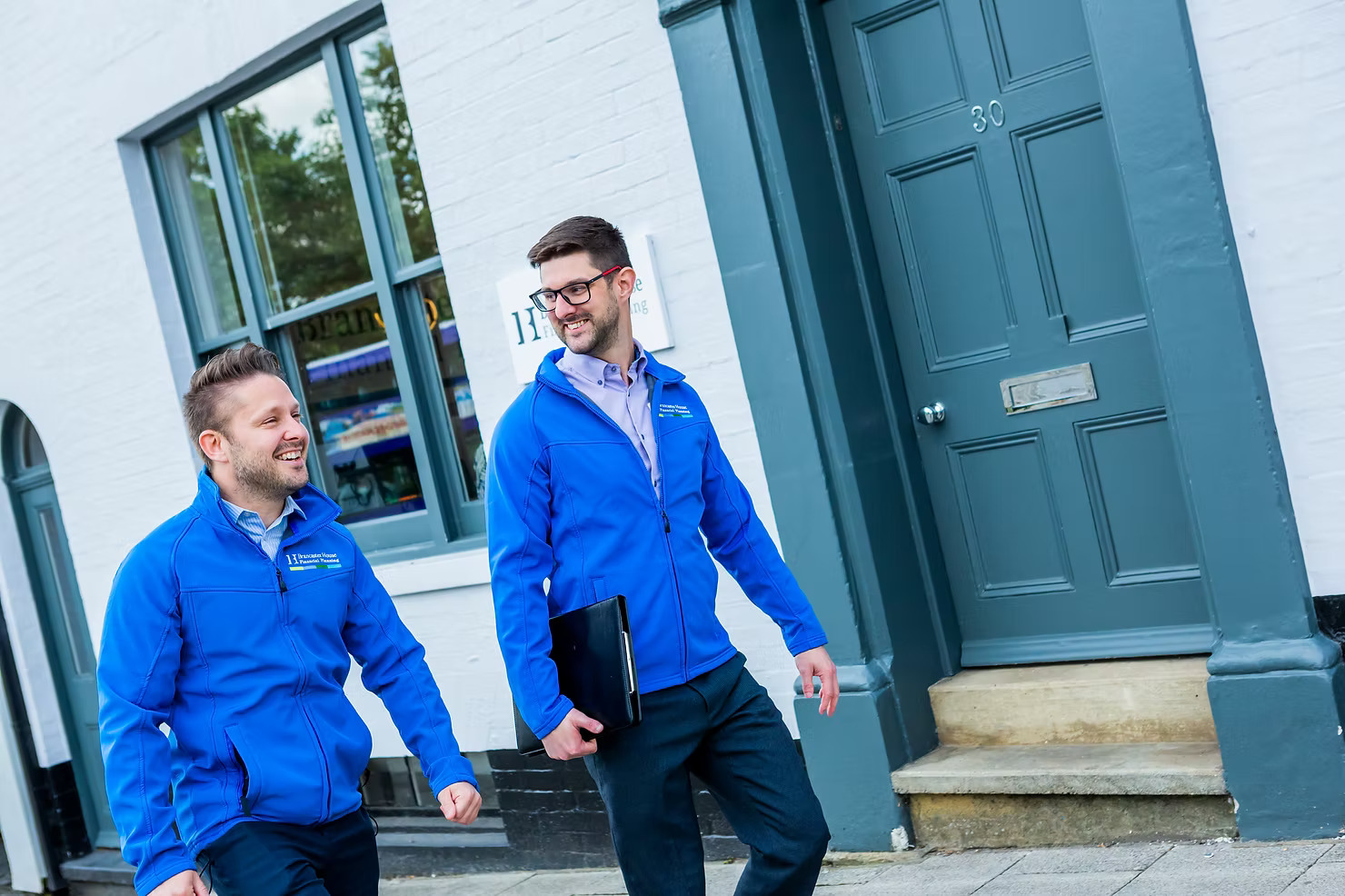 Two men in blue jackets walk and chat happily outside a white building with teal doors and windows. One carries a black folder.